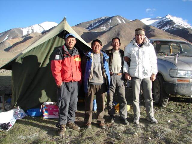 Four men in front of a tent and a vehicle. They are smiling toward the camera.