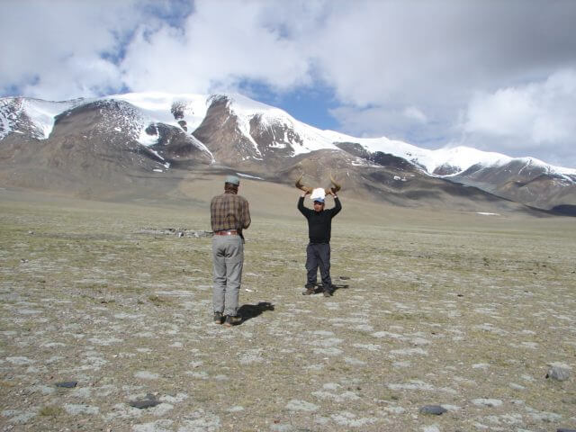 Two men stand near a mountain. The one facing the camera is holding a yak skull above his head by the horns.