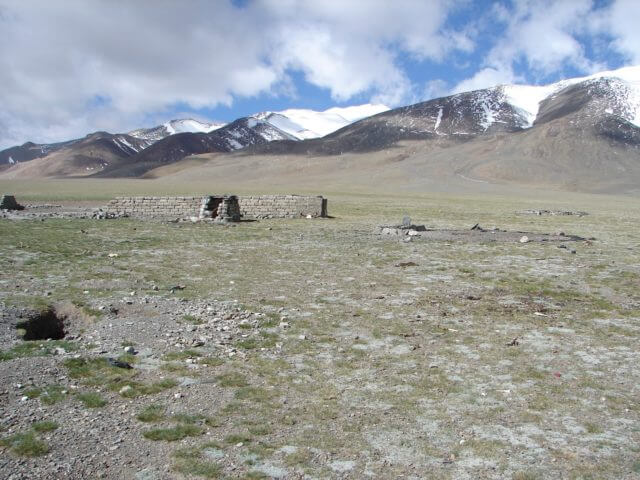 Several crumbling low brick walls sit near a circle of bare earth ringed with stones, snowy mountains behind.