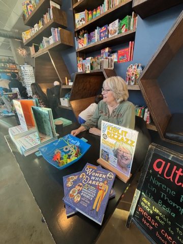 Author Linda Skeers sitting at a table during a book event with her books displayed nearby